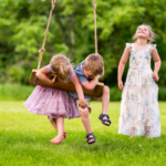 young girl and boy hang from an old-fashioned swing while another child looks up at the sky in a family photo