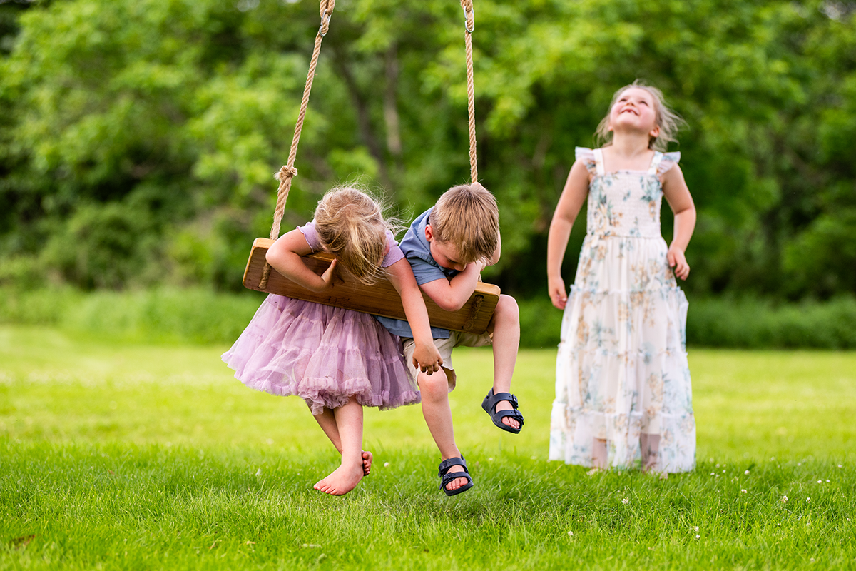 young girl and boy hang from an old-fashioned swing while another child looks up at the sky in a family photo
